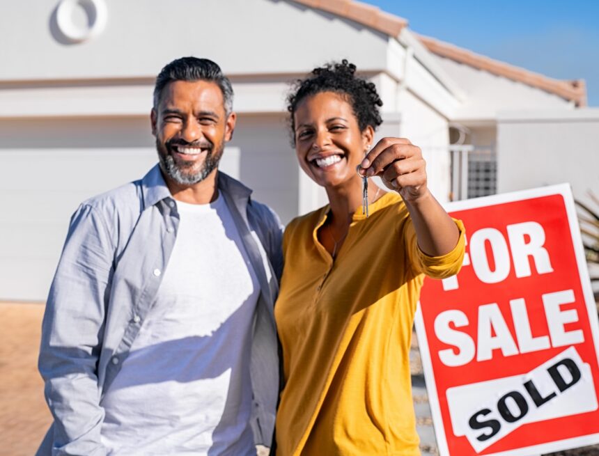 A couple stands outside a home they just bought, with a woman holding keys