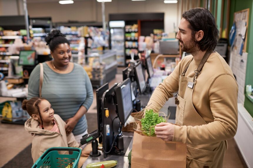 smiling-male-worker-in-supermarket-helping-young-w-2025-03-05-11-32-27-utc A woman checks out at a grocery store with her daughter