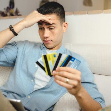young-hispanic-man-with-a-worried-face-doing-onlin-2025-02-02-15-02-14-utc A shallow focus of a young Hispanic man with a worried face sitting on the sofa at home holding many credit cards