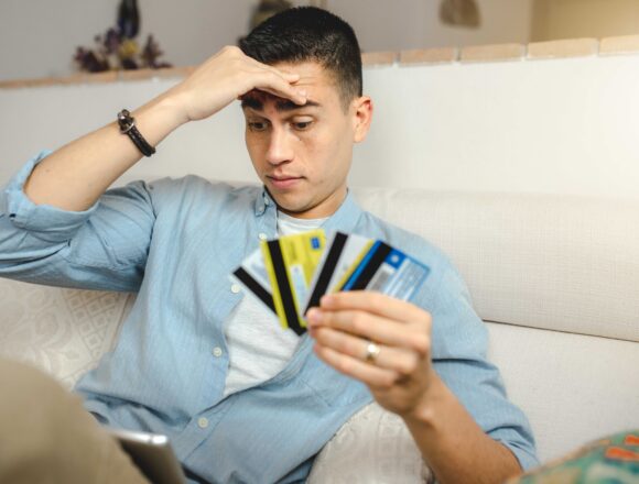 A shallow focus of a young Hispanic man with a worried face sitting on the sofa at home holding many credit cards